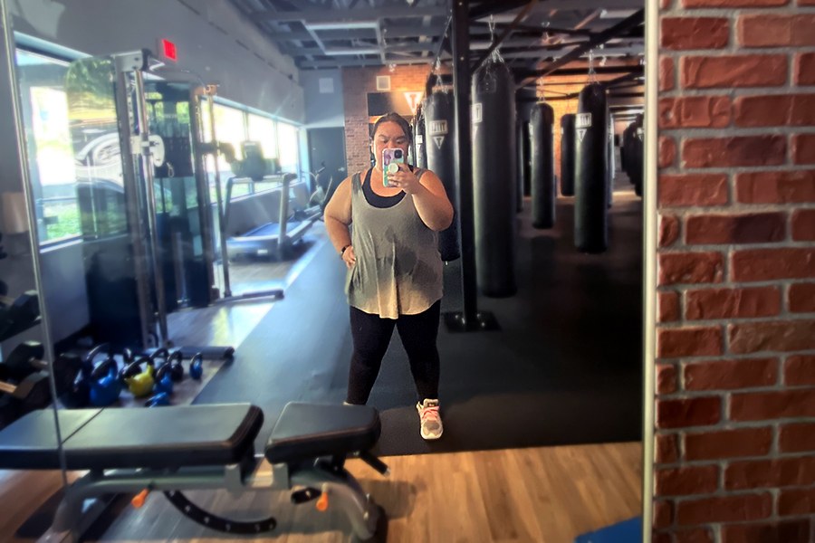 Coach Rose standing in front of a mirror at the end of a boxing class. Black heavy bags are in the background of the gym